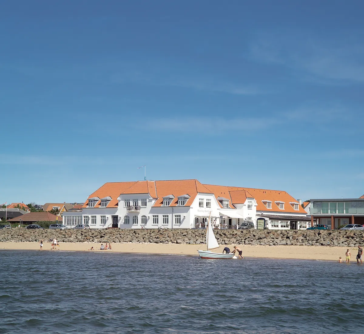 Hjerting Badehotel set fra vandet med strandliv og en sejlbåd i forgrunden